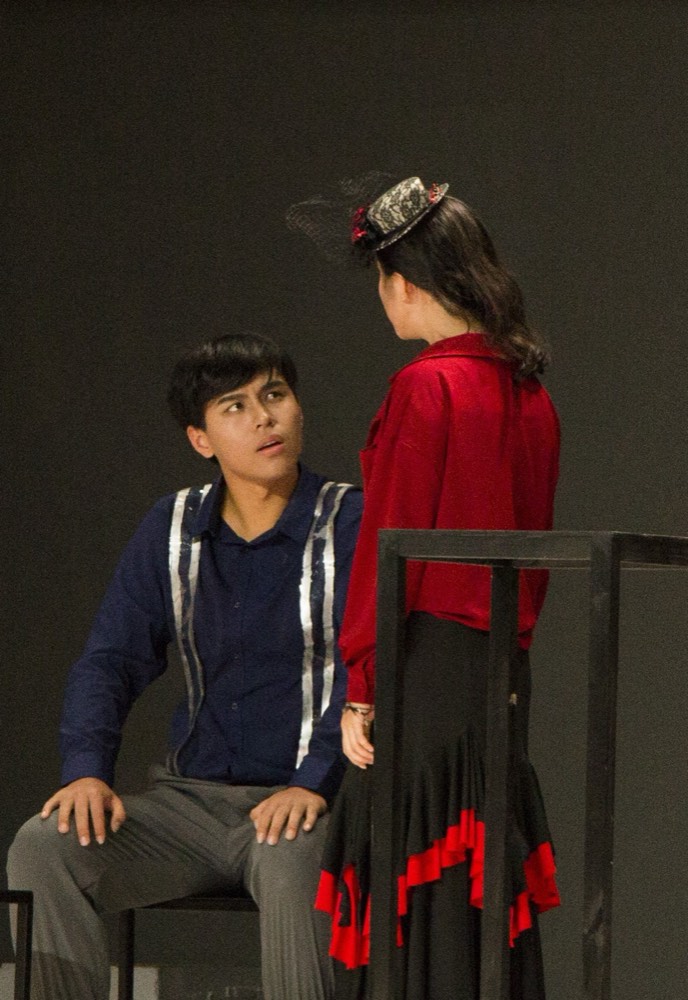 Two actors in a scene — one seated, looking up at the other, who stands over him in red.