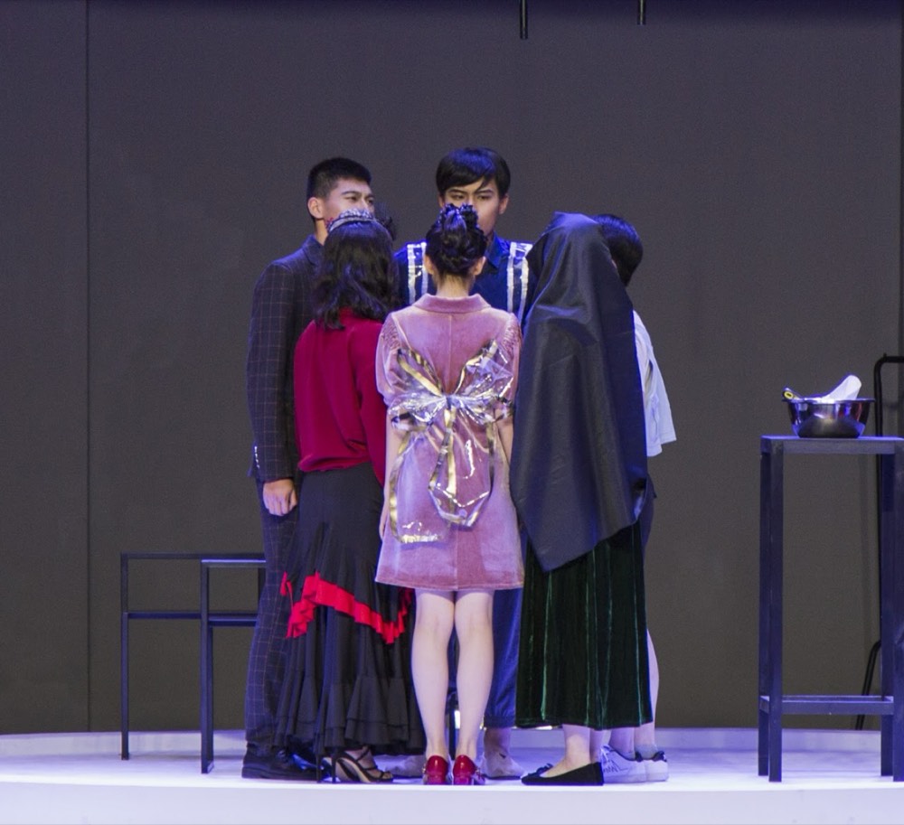 A tight cluster of actors seen from behind, gathered centre stage against a dark wall.