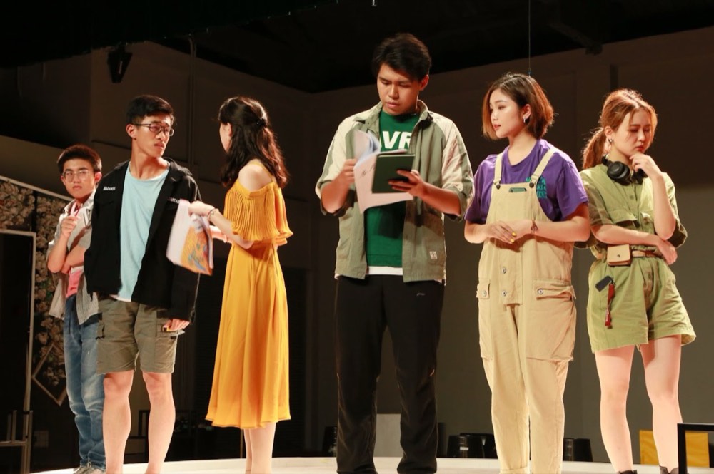 Six actors in a line mid-scene, two of them reading from papers.